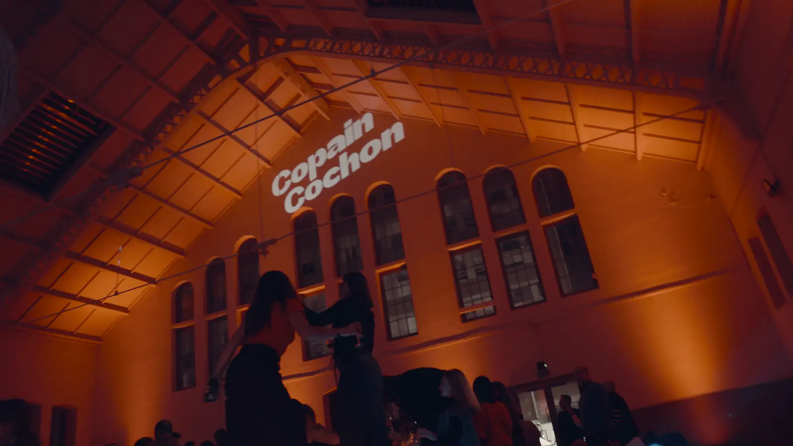 Convives qui dansent dans une halle éclairée en orange, sous le logo Copain Cochon